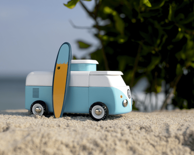 Toy van with a surfboard on a sandy surface with greenery in the background